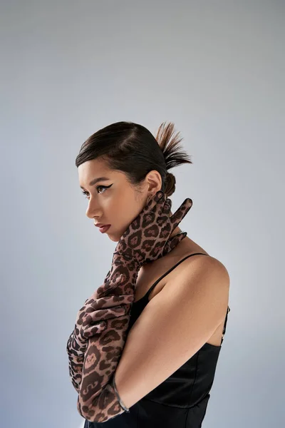 Graceful asian woman with trendy hairstyle and bold makeup posing in black strap dress and animal print gloves while looking away on grey background, spring fashion photography — Stock Photo