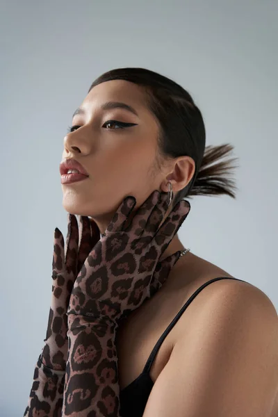 Youthful style, spring fashion, young asian woman with brunette hair and bold makeup, wearing black dress and animal print gloves, touching neck, expressive, on grey background — Stock Photo