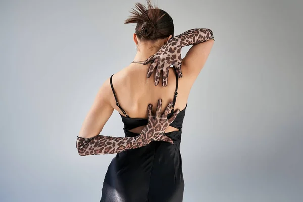 Back view of young brunette woman with trendy hairstyle posing with hands behind back on grey background, black strap dress, animal print gloves, trendy spring, fashion photography — Stock Photo