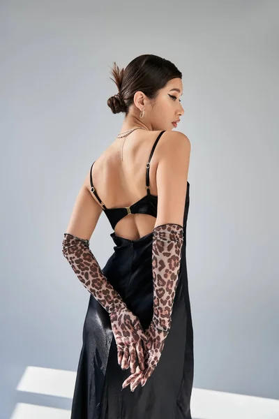 Back view of asian fashion model with brunette hair standing in black strap dress and animal print gloves on grey background with lighting, youth culture, spring fashion, generation z — Stock Photo