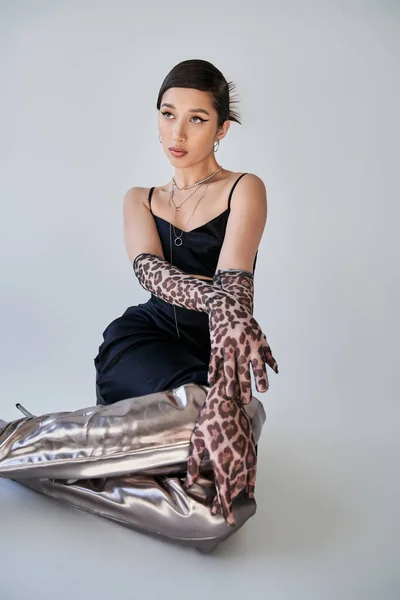 Элегантная азиатская женщина с смелым макияжем, в черном ремешке платье, перчатки для печати животных и серебряные сапоги сидя и глядя в сторону на сером фоне, молодой стиль, модная весенняя концепция — стоковое фото