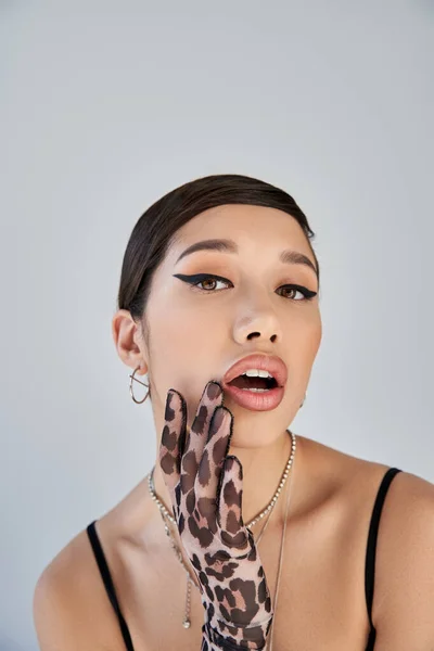Портрет соблазнительной азиатской женщины с смелым макияжем, в серебряных ожерельях и перчатках для печати животных, держащей руку у открытого рта и смотрящей на камеру на сером фоне, весенняя концепция моды — стоковое фото