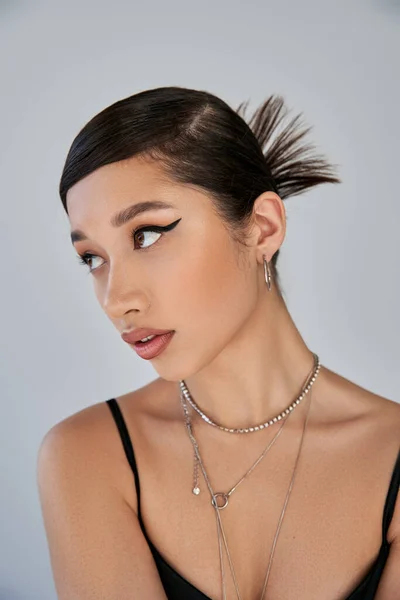 Portrait of attractive asian woman with brunette hair, expressive gaze, bold makeup, trendy hairstyle and silver necklaces looking away on grey background, youthful style, trendy spring concept — Stock Photo