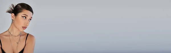 Porträt einer charmanten Asiatin mit trendiger Frisur und kühnem Make-up auf grauem Hintergrund, schwarzem Trägerkleid, silbernen Accessoires, ausdrucksstarkem Blick, Frühjahrsmodekonzept, Banner — Stockfoto
