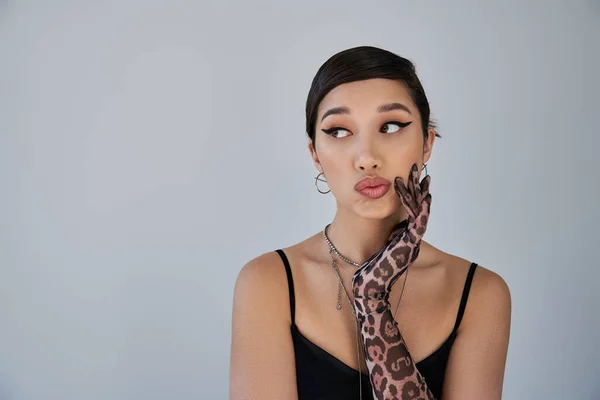 Portrait of fashionable asian woman with skeptical face expression looking away on grey background, brunette hair, silver accessories, animal print glove, black dress, trendy spring concept — Stock Photo