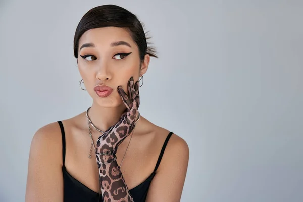 Portrait of young asian woman in black strap dress, silver necklaces and animal print glove, with thoughtful and skeptical face expression looking away on grey background, spring fashion photography — Stock Photo