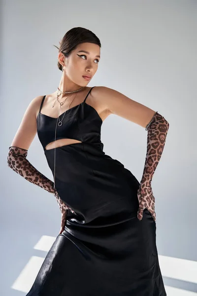 Fashionable asian woman with bold makeup and brunette hair holding hands on hips while posing in black strap dress and animal print gloves on grey background, trendy spring, generation z — Stock Photo
