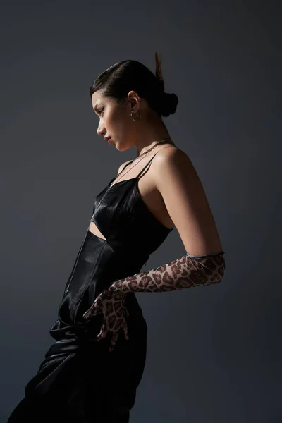 Vista lateral de mujer asiática joven y bonita con peinado de moda posando con la mano en la cadera en vestido de correa negro y guante de impresión animal sobre fondo gris oscuro, concepto de primavera de moda - foto de stock
