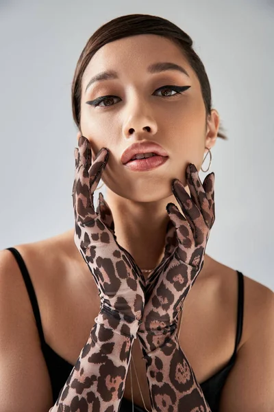 Portrait of youthful and beautiful asian fashion model with brunette hair and bold makeup posing in animal print gloves and looking at camera on grey background, trendy spring concept — Stock Photo