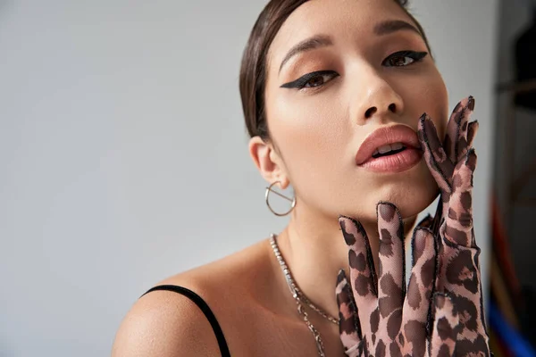Portrait of expressive asian woman with bold makeup, brunette hair and seductive gaze touching face and looking at camera on grey background, silver accessories, animal print gloves, stylish spring — Stock Photo