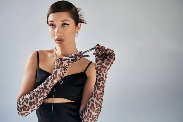 Stunning asian woman with bold makeup and trendy hairstyle, wearing silver necklaces and black strap dress, taking off animal print glove and looking away on grey background, trendy spring — Stock Photo