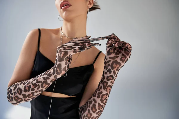 Cropped view of young sensual woman in black strap dress and silver accessories taking off stylish animal print glove on grey background, generation z, trendy fashion concept — Stock Photo