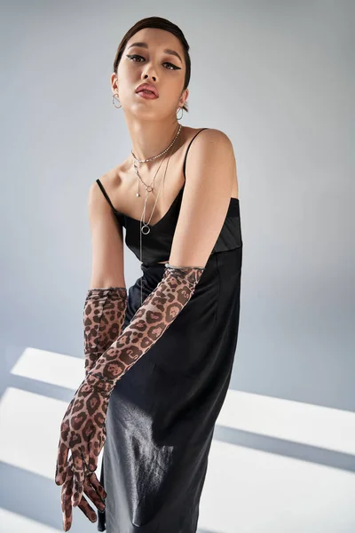 Spring fashion, seductive asian woman with expressive gaze and bold makeup looking at camera on grey background with lighting, black strap dress, animal print gloves, silver necklaces — Stock Photo