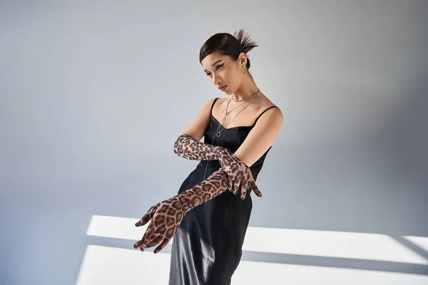 Youthful asian fashion model with trendy hairstyle and bold makeup posing in trendy spring outfit, black strap dress and animal print gloves on grey background with lighting — Stock Photo