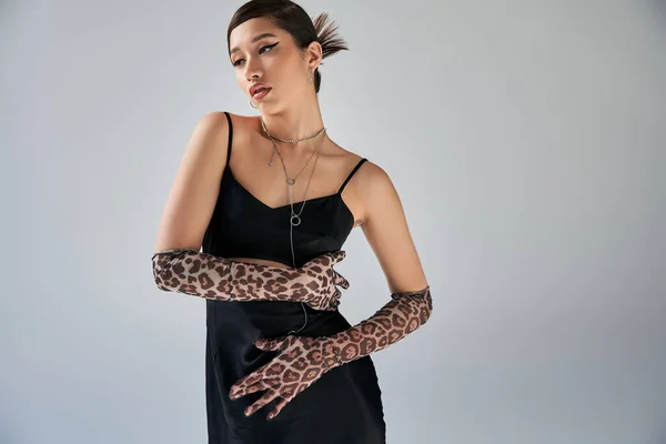 Attracive asian woman with bold makeup and brunette hair posing in silver necklaces, black strap dress and animal print gloves on grey background, high fashion, spring style — Stock Photo