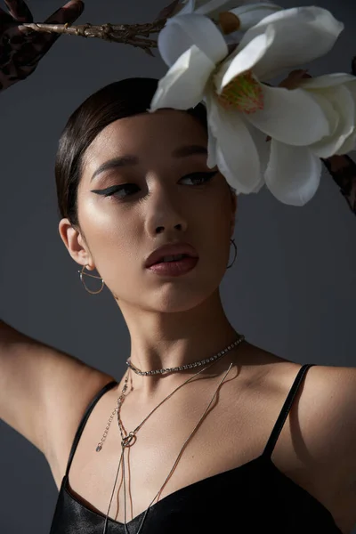 Portrait of expressive asian woman in silver necklaces and black strap dress, with brunette hair and bold makeup holding white orchid on dark grey background, fashionable spring concept — Stock Photo