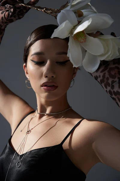 Portrait of young asian fashion model with bold makeup and brunette hair wearing black strap dress and silver necklaces while posing with white orchid on dark grey background, spring style concept — Stock Photo