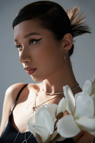 Весенний стиль, портрет привлекательной азиатской женщины с брюнетками, серебряные ожерелья, смелый макияж и модная прическа глядя в сторону белой орхидеи на сером фоне, поколение z — стоковое фото