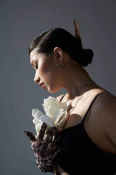 Привлекательная азиатская женщина с брюнетками, смелым макияжем и серебряными аксессуарами, в черном платье и перчатке для печати животных с белой орхидеей на темно-сером фоне, стильная весенняя концепция — стоковое фото
