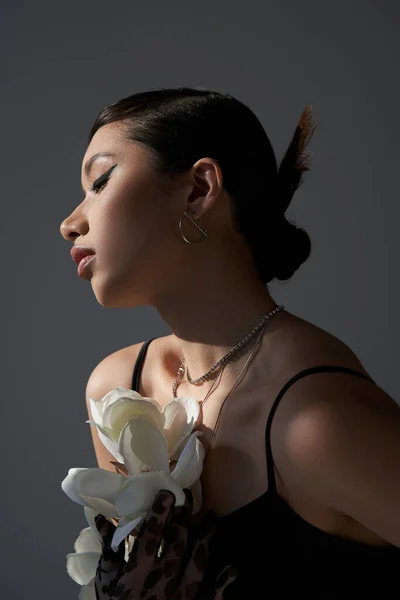 Fotografía de moda de primavera, atractiva mujer asiática con maquillaje audaz y peinado de moda, en collares de plata y vestido de correa negro posando con orquídea blanca sobre fondo gris oscuro, generación z - foto de stock