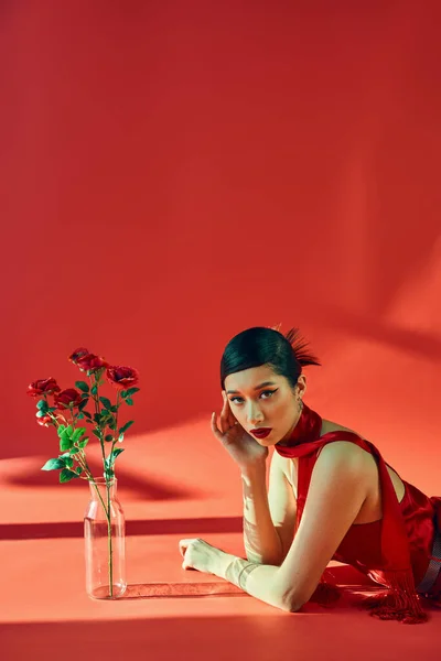 Concepto de moda de primavera, mujer asiática joven con cabello moreno y maquillaje audaz que pone en el pañuelo y el vestido mientras mira a la cámara cerca de rosas frescas sobre fondo rojo con iluminación - foto de stock