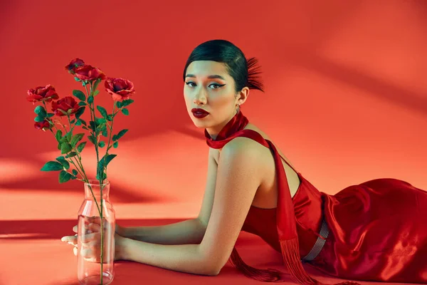 Concepto de moda de primavera, mujer asiática joven y elegante con maquillaje audaz, en el pañuelo y el vestido acostado cerca de jarrón de cristal con rosas y mirando a la cámara en el fondo rojo con iluminación - foto de stock
