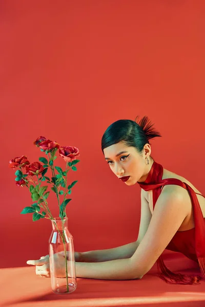 Young and elegant asian woman in stylish neckerchief and dress, with bold makeup and brunette hair laying near glass vase with roses on red background with lighting, spring fashion, generation z — Stock Photo