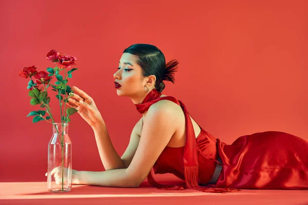Side view of charming asian woman with brunette hair and bold makeup, in elegant dress and neckerchief lying and touching roses in glass vase on red background with lighting, spring fashion concept — Stock Photo