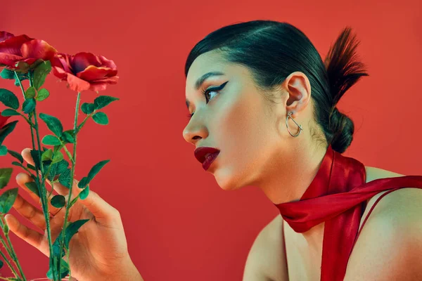 Боковой вид завораживающей азиатской женщины с брюнетками, смелым макияжем и модной прической, касающейся зеленых листьев роз на красном фоне, поколение z, весенняя концепция моды — стоковое фото