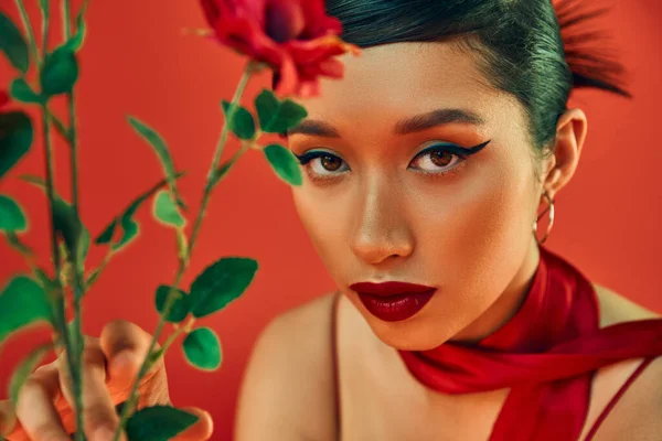 Portrait of young asian woman in neckerchief, with bold makeup and expressive gaze looking at camera near flowers on red background, spring fashion photography, generation z — Stock Photo