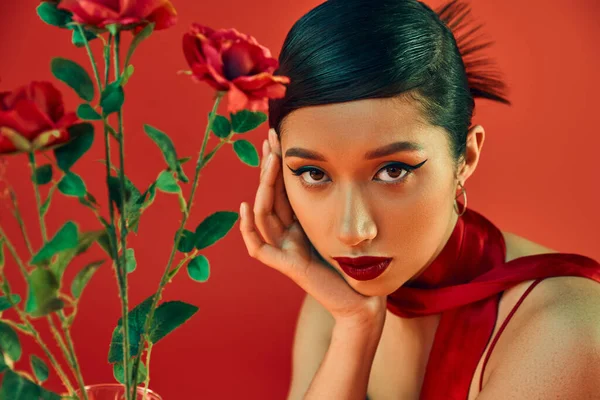 Retrato de atractiva mujer asiática con maquillaje audaz, cabello moreno y mirada expresiva mirando a la cámara cerca de rosas sobre fondo rojo, primavera de moda, estilo juvenil - foto de stock
