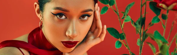 Портрет молодой и заманчивой азиатской женщины с смелым макияжем и выразительным взглядом, держащимся за руку рядом с лицом и смотрящей в камеру рядом с цветами на красном фоне, модной весной, поколением z, баннером — стоковое фото