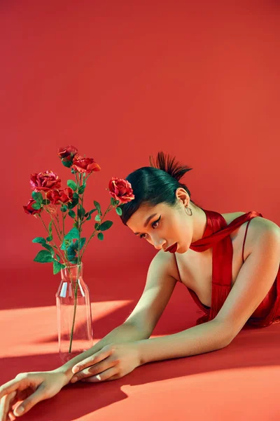 Encantadora mujer asiática con cabello moreno y maquillaje audaz con pañuelo y puesta sobre fondo rojo con iluminación cerca de jarrón de cristal con rosas, moda juvenil, primavera de moda - foto de stock