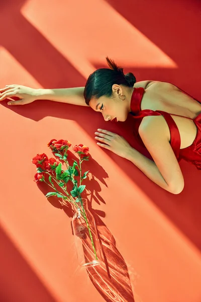 Вид на выразительную и молодую азиатскую женщину с брюнетками, в шейном платке и платье, лежащее в освещении возле стеклянной вазы с розами на красном фоне, поколение z, весенняя модная фотография — стоковое фото