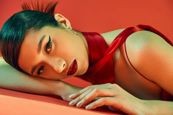 Femme asiatique sensuelle et charmante avec un regard expressif posé sur fond rouge et regardant la caméra, maquillage audacieux, cheveux bruns, foulard, photographie de mode de printemps — Photo de stock