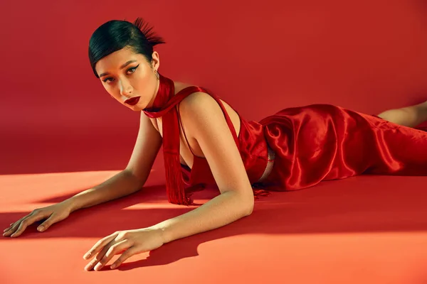 Спокуслива азіатська жінка з модною зачіскою і сміливим макіяжем, в елегантному платті і хустці лежить і дивиться на камеру на червоному тлі з освітленням, молодіжною культурою, стильною концепцією весни — стокове фото