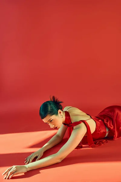 Femme asiatique sensuelle avec coiffure tendance et maquillage audacieux, en foulard et robe élégante posant sur fond rouge avec éclairage, concept printemps tendance, génération z — Photo de stock