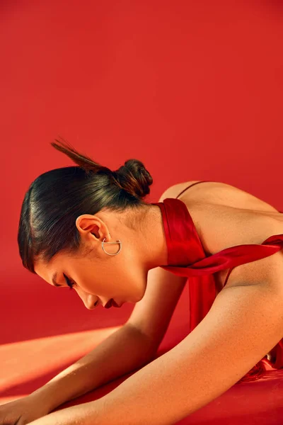 Вид сбоку молодой и чувственной азиатской женщины с брюнетками, смелый макияж, модная прическа в серебряной сережке и шейный платок на красном фоне с освещением, модная весенняя концепция — стоковое фото