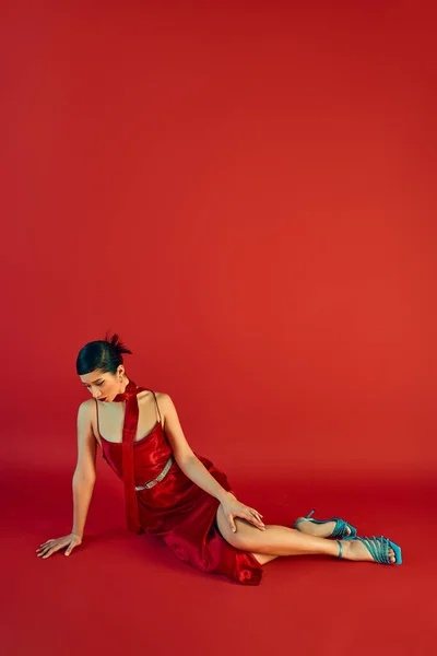 Frühling Eleganz, volle Länge der jungen asiatischen Frau in stilvollem Kleid, Halstuch und türkisfarbenen Sandalen sitzen auf rotem Hintergrund mit Kopierraum, Generation z, Mode-Shooting — Stockfoto