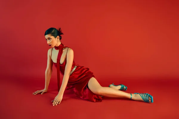 Moda de primavera, longitud completa de la mujer asiática de moda con cabello moreno y maquillaje audaz mirando hacia otro lado mientras está sentado sobre fondo rojo en vestido de correa elegante, sandalias de color turquesa y pañuelo - foto de stock