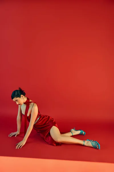Piena lunghezza di giovane e graziosa donna asiatica in abito cinturino, sandali turchese e fazzoletto seduto in posa espressiva su sfondo rosso, gen z moda, abito primaverile — Foto stock