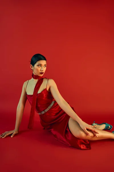 Очаровательная азиатская женщина с брюнеткой волосы и смелый макияж глядя в сторону, сидя на красном фоне в галстуке и ремешок платье, поколение z, весенняя концепция моды — стоковое фото