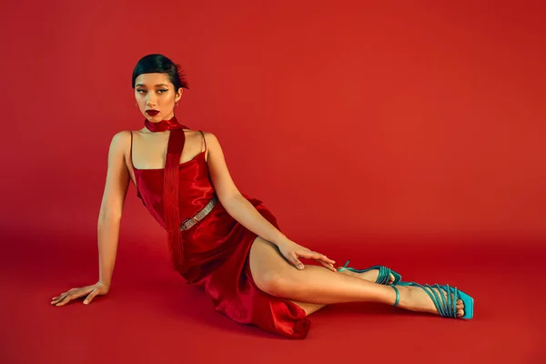 Trendy spring concept, full length of mesmerizing asian fashion model with brunette hair and bold makeup, in strap dress, neckerchief and turquoise sandals sitting and looking away on red background — Stock Photo