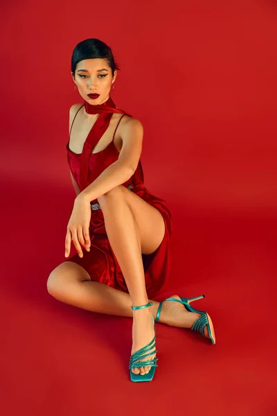 Pleine longueur de femme asiatique élégante en tenue de printemps élégant assis dans une pose élégante sur fond rouge, cheveux bruns, robe de sangle, foulard, sandales turquoise, génération z mode — Photo de stock