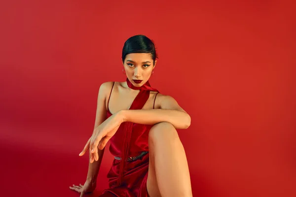 Conceito de primavera elegante, gen z moda, jovem mulher asiática com cabelo morena e maquiagem ousada sentado em vestido de cinta e cachecol enquanto olha para a câmera no fundo vermelho — Fotografia de Stock