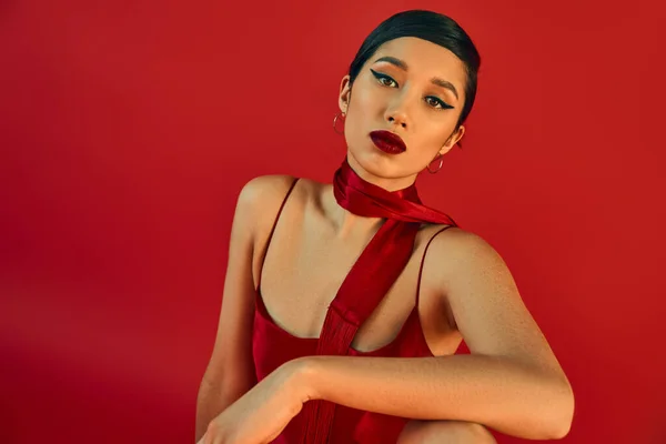 Gen z fashion, spring style, portrait of young and beautiful asian woman with bold makeup and expressive gaze looking at camera while posing in strap dress and neckerchief on red background — Stock Photo