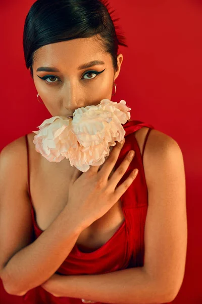 Portrait of young and sensual asian woman with brunette hair and bold makeup, in neckerchief and strap dress posing with white peony on vibrant red background, trendy spring concept — Stock Photo