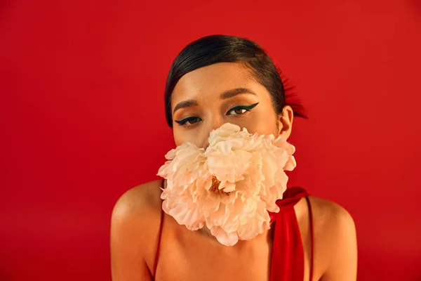 Portrait de jeune et sensuel modèle asiatique avec maquillage audacieux et cheveux bruns tenant pivoine blanche dans la bouche et regardant la caméra sur fond rouge vif, gen z mode, style printemps — Photo de stock