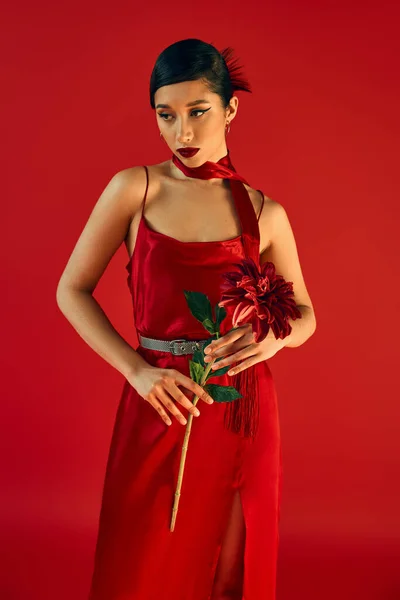 Jolie et à la mode asiatique femme en robe à bretelles noires et foulard, avec cheveux bruns et maquillage audacieux debout avec pivoine bordeaux sur fond rouge — Photo de stock