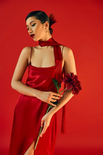 Молодая и чувственная азиатская женщина с смелым макияжем и брюнетками, в гламурном платье и с галстуком, позирующая с бордовым пионом на красном фоне, модная весна, ген з мода — стоковое фото